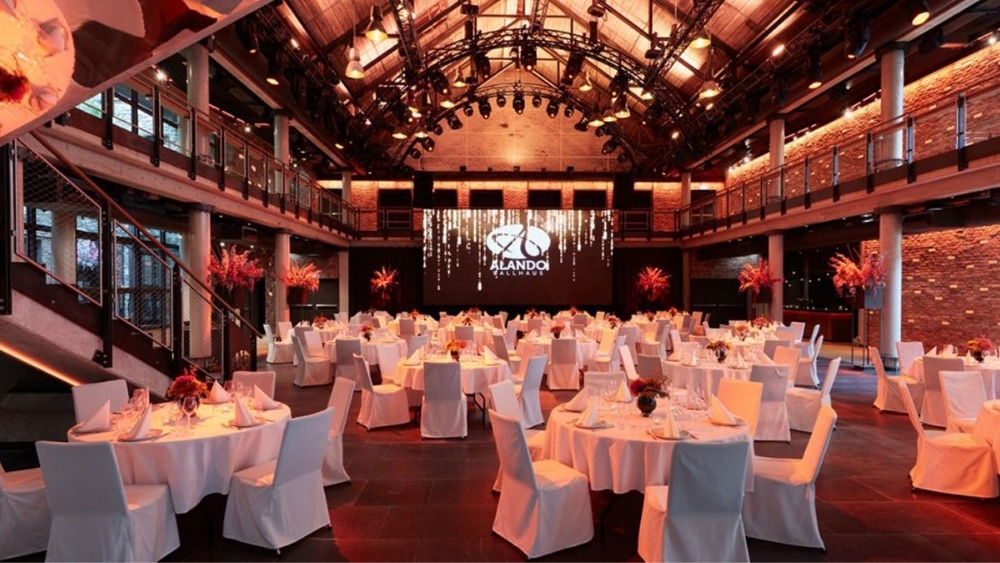 Moderne Veranstaltungshalle mit festlich gedeckten Tischen und Bühnenpräsentation, ideal für Events.
