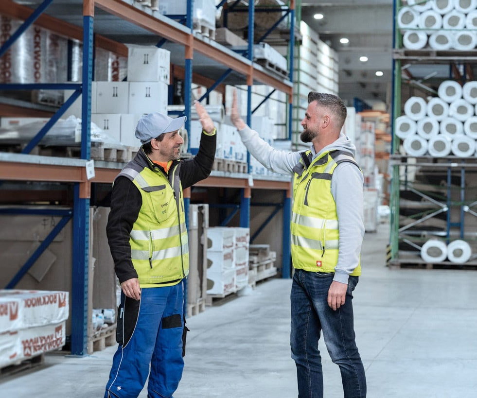 Zwei Mitarbeiter in Baustofflager, high-five, mit Regalen voller Baumaterialien im Hintergrund.
