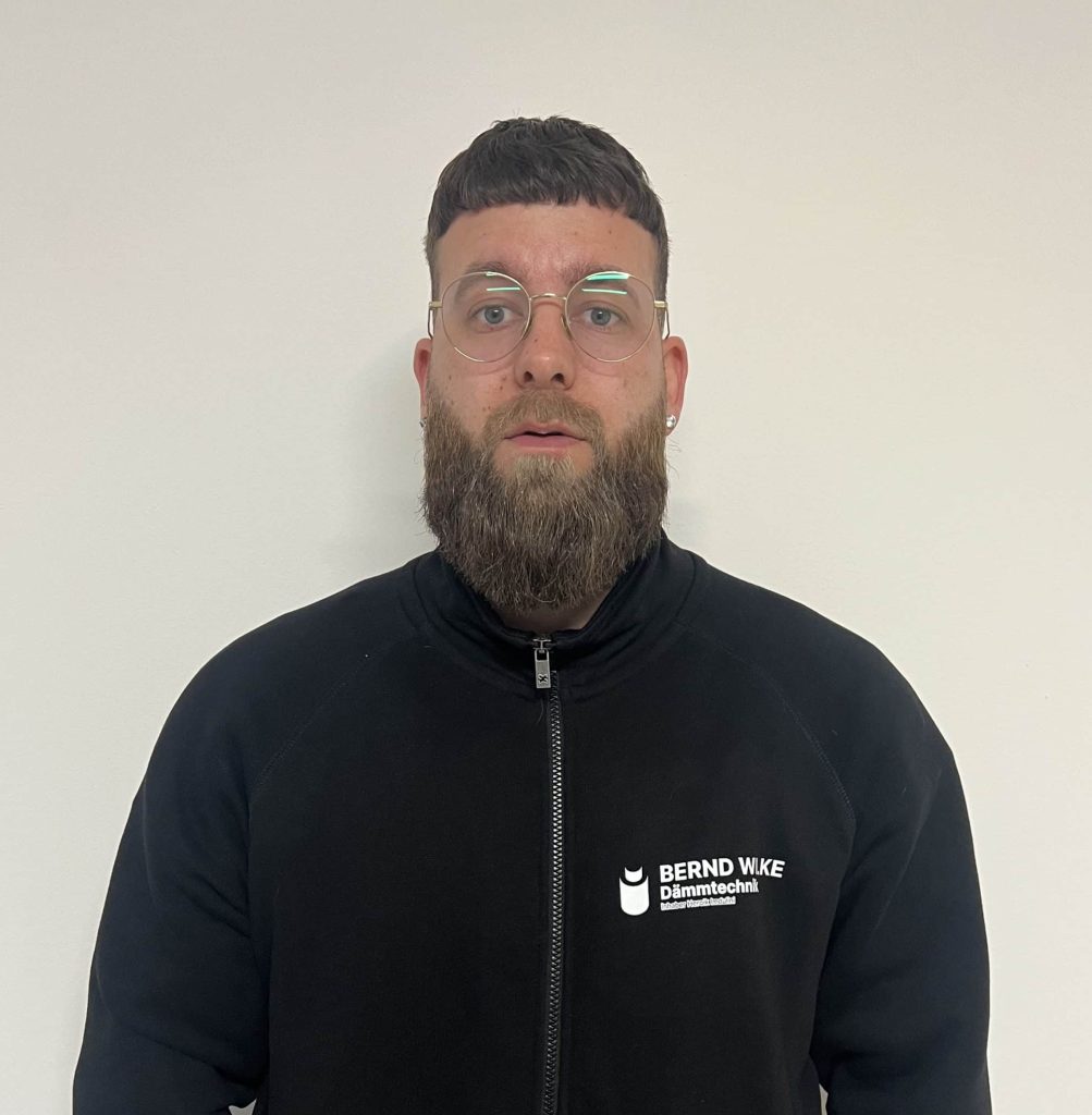 Herr in schwarzem Wego Systembaustoffe Pullover vor weißer Wand, professionell und freundlich.